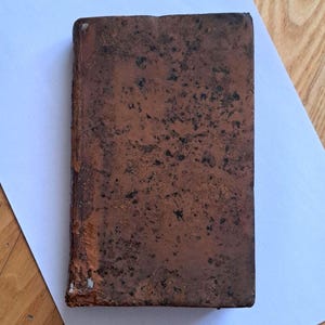 May include: An antique book with a worn, brown cover. The cover has a textured appearance with dark spots and signs of wear on the edges. The book is resting on a white sheet of paper.
