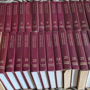 May include: A collection of thirty maroon hardback books arranged in two rows. Each spine features gold lettering with the title Bolshaya Sovetskaya Entsiklopediya and a unique volume number ranging from one to thirty, with two volume 24s.