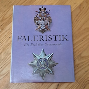 May include: A book titled "Faleristik" with the subtitle "Ein Buch über Ordenskunde" on a purple background. The book features images of medals and awards. The book is lying on a wooden surface.
