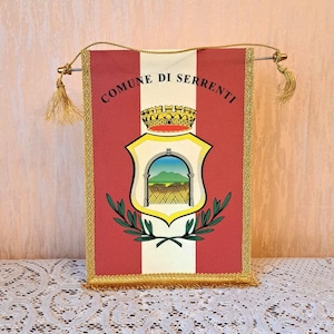 Könnte beinhalten: Ein rechteckiges Banner mit dem Text "COMUNE DI SERRENTI" und einem Wappen. Das Banner ist rot mit goldenen Verzierungen und Quasten. Das Wappen zeigt einen Schild mit einer Landschaftsszene und einer Krone.