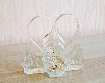 May include: A clear glass ring holder shaped like two swans with their necks intertwined, forming a heart shape. Two gold rings are nestled in the center. The holder sits on a light wooden surface.