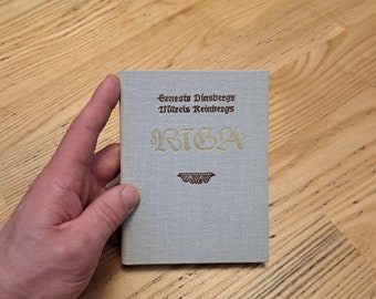 May include: A blue book with the title "RIGA" in gold lettering. The book is by authors "Ernests Dinsbergs" and "Māris Reinbergs".