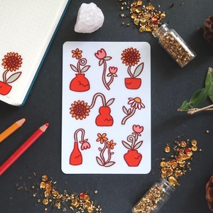 May include: A sheet of floral stickers featuring various flowers in orange pots and vases. The stickers have a retro design with a white background. A matching sticker is on a notebook. Gold sequins and pencils are scattered around the stickers.