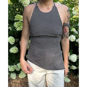 May include: A black and white checkered halter top with a fitted bodice. The top is paired with cream-colored pants. The background features green foliage and white flowers.