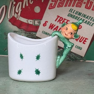 May include: A white ceramic mug with green holly leaf designs. The handle is a green elf figurine. The background features a vintage Santa Claus advertisement with the words "Santa-Glo" and "Illuminated Unbreakable".