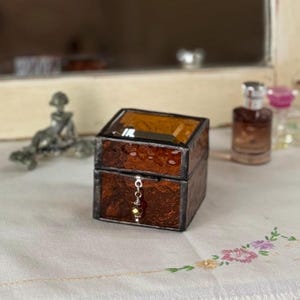 Puede incluir: Una pequeña caja cuadrada de vidrio marrón con un interior dorado. La caja tiene una bisagra de metal y un pequeño amuleto dorado colgando.