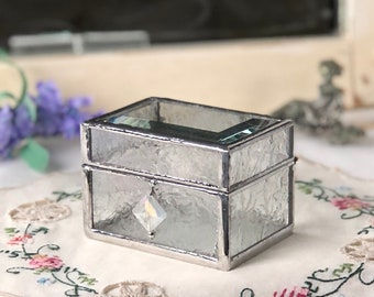 May include: A small, clear glass ring box with a frosted finish and a silver metal frame. The box has a clear crystal on the front.