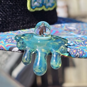 May include: A unique, clear glass art piece with a blue-green hue, featuring a small, enclosed figure. The glass has a dripped design with rounded edges. The piece is set on a patterned surface, with a black hat in the background.