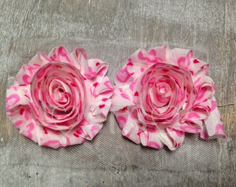 May include: Two pink fabric flowers with a white background and pink hearts printed on them. The flowers are attached to a piece of white tulle.
