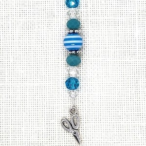 May include: A silver charm with a blue and white striped bead and blue crystal beads. The charm is attached to a silver lobster clasp. The charm is hanging from a silver chain with a pair of silver scissors at the bottom.