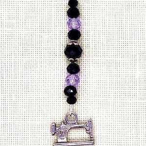 May include: A silver-toned charm with a sewing machine pendant. The charm features a strand of black and purple beads, and a silver clasp. This item is perfect for sewing enthusiasts.