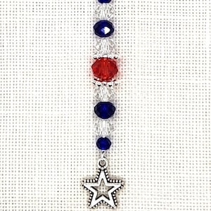 May include: A silver charm with a star pendant and a beaded chain in red, white, and blue colors. The charm has a silver lobster clasp.