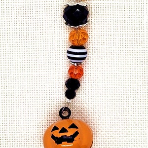 May include: A Halloween-themed charm with a pumpkin-shaped bell and a beaded chain featuring black, orange, and white beads. The chain has a silver clasp.