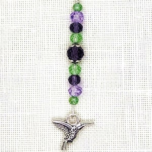 May include: A silver hummingbird charm hangs from a silver chain with green, purple, and black crystal beads.