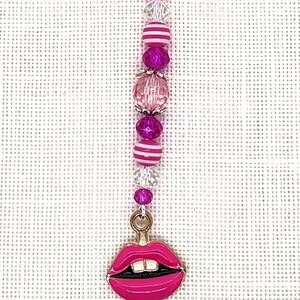May include: A pink and white striped charm with a pink lip charm attached to a silver clasp. The charm is made of beads and is approximately 6 inches long.