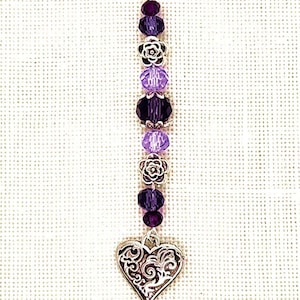 May include: A silver charm with a purple and lavender crystal bead and flower accents. The charm is attached to a silver lobster clasp.