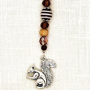 May include: A beaded charm with a silver squirrel pendant. The charm features a series of beads in shades of brown, purple, and white, including wooden and striped beads. The charm is attached to a long, thin metal pin.