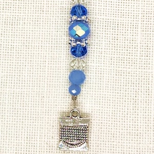 May include: A blue and silver beaded needle minder with a small silver spool charm. The needle minder is strung on a thin wire with a silver loop at the top.