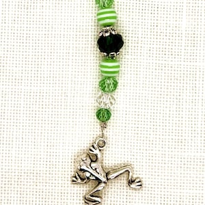 May include: A silver frog charm hangs from a beaded chain with green and white striped beads and clear crystal beads. The chain is attached to a silver pin.
