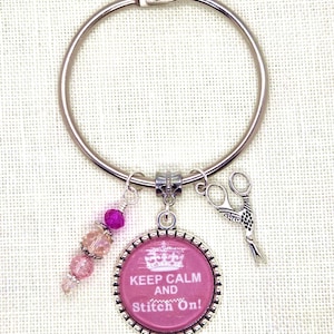 May include: A silver-tone keychain with a pink charm that reads "Keep Calm and Stitch On!" along with a small pair of scissors and a beaded charm. The keychain is designed for sewing enthusiasts.