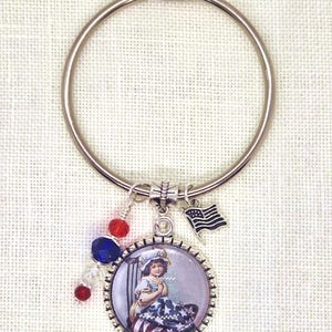 May include: A silver-tone keychain with a patriotic theme. It features a round pendant with a vintage image of a child, a small American flag charm, and red and blue beads. The keychain is designed to celebrate American pride.