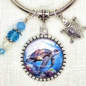 K&ouml;nnte beinhalten: Ein silberfarbenes Armband mit einem Anh&auml;nger in Form einer Meeresschildkr&ouml;te. Der Anh&auml;nger zeigt ein detailliertes Bild einer Meeresschildkr&ouml;te, die im Ozean schwimmt. Zus&auml;tzliche Charms sind eine kleine Schildkr&ouml;te und blaue und klare Perlen.