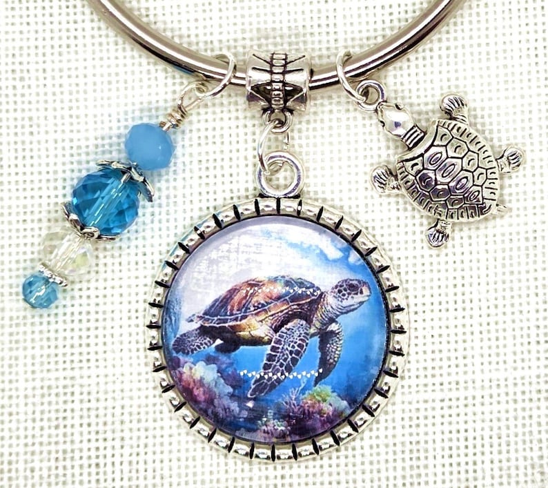 K&ouml;nnte beinhalten: Ein silberfarbenes Armband mit einem Anh&auml;nger in Form einer Meeresschildkr&ouml;te. Der Anh&auml;nger zeigt ein detailliertes Bild einer Meeresschildkr&ouml;te, die im Ozean schwimmt. Zus&auml;tzliche Charms sind eine kleine Schildkr&ouml;te und blaue und klare Perlen.