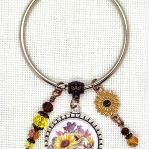 May include: A silver metal keyring with a floral charm. The charm features a sunflower design with yellow, orange, and brown accents. Additional charms include a yellow and brown beaded strand and a small sunflower pendant.