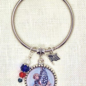 May include: A silver-tone keychain with a round pendant featuring a vintage illustration of a child holding an American flag. The keychain includes a small flag charm and colorful beads, perfect for patriotic themes.