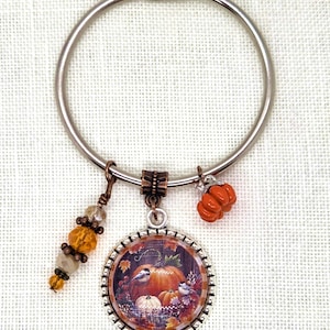May include: A silver-tone keychain with fall-themed charms. The main charm is a round pendant with a pumpkin illustration. Additional charms include an orange pumpkin, and a beaded accent with amber and clear beads.
