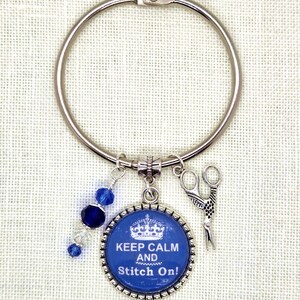 Puede incluir: Un anillo plateado con un dije azul y blanco que dice "Keep Calm and Stitch On!". El dije presenta un dise&ntilde;o de corona y est&aacute; acompa&ntilde;ado de un dije de tijeras y cuentas azules.