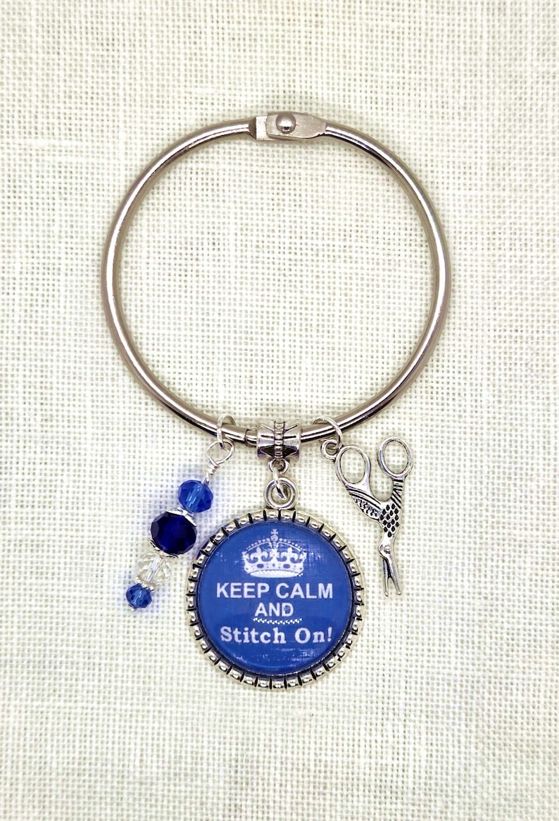 Puede incluir: Un anillo plateado con un dije azul y blanco que dice "Keep Calm and Stitch On!". El dije presenta un dise&ntilde;o de corona y est&aacute; acompa&ntilde;ado de un dije de tijeras y cuentas azules.