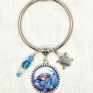 K&ouml;nnte beinhalten: Ein silberfarbener Schl&uuml;sselanh&auml;nger mit Meeresschildkr&ouml;ten-Anh&auml;ngern. Der Schl&uuml;sselanh&auml;nger hat einen runden Anh&auml;nger mit einem Meeresschildkr&ouml;ten-Bild, einen kleinen Schildkr&ouml;ten-Anh&auml;nger und blaue Perlenakzente. Der Hintergrund ist ein heller Stoff.