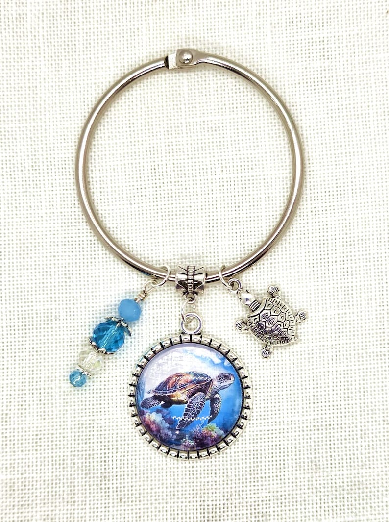 K&ouml;nnte beinhalten: Ein silberfarbener Schl&uuml;sselanh&auml;nger mit Meeresschildkr&ouml;ten-Anh&auml;ngern. Der Schl&uuml;sselanh&auml;nger hat einen runden Anh&auml;nger mit einem Meeresschildkr&ouml;ten-Bild, einen kleinen Schildkr&ouml;ten-Anh&auml;nger und blaue Perlenakzente. Der Hintergrund ist ein heller Stoff.