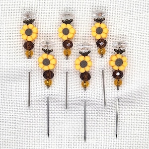 May include: A collection of decorative sewing pins. Each pin features a yellow sunflower design with a brown center, a clear crystal top, and brown and yellow beads. The pins are arranged on a white fabric background.