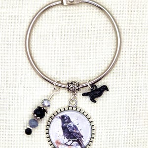 May include: A silver-tone keychain with a raven theme. It features a round pendant with a raven illustration, a small black raven charm, and a strand of black and gray beads. The keychain is designed for keys or bags.