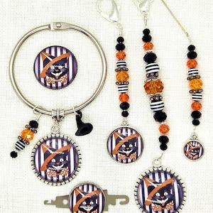 May include: A collection of Halloween-themed accessories featuring a black cat wearing a witch hat and bow tie. The set includes a key ring, charms, and beaded accents in black, orange, and white. The cat design is set against a striped background.
