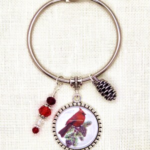 May include: A silver-tone keyring with a red cardinal bird pendant, a pinecone charm, and a beaded dangle. The cardinal is perched on a pine branch. The beads are red and clear.