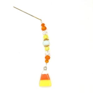 May include: A decorative charm featuring a vertical arrangement of orange, clear, and yellow faceted beads strung on a thin metal wire, terminating in a triangular candy corn pendant with white, orange, and yellow horizontal color bands.