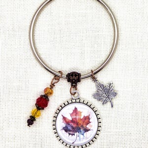 May include: A silver key ring with three charms. One charm is a round pendant with a watercolor maple leaf design. Another is a silver maple leaf charm. The third charm is a cluster of colorful beads.