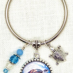 K&ouml;nnte beinhalten: Ein silberfarbener Schl&uuml;sselanh&auml;nger mit einem Anh&auml;nger in Form einer Meeresschildkr&ouml;te, einem kleinen Schildkr&ouml;ten-Charm und blauen Perlenakzenten. Der Anh&auml;nger zeigt eine detaillierte Abbildung einer Meeresschildkr&ouml;te im Meer. Ein dekoratives Accessoire.