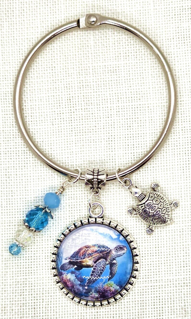 K&ouml;nnte beinhalten: Ein silberfarbener Schl&uuml;sselanh&auml;nger mit einem Anh&auml;nger in Form einer Meeresschildkr&ouml;te, einem kleinen Schildkr&ouml;ten-Charm und blauen Perlenakzenten. Der Anh&auml;nger zeigt eine detaillierte Abbildung einer Meeresschildkr&ouml;te im Meer. Ein dekoratives Accessoire.