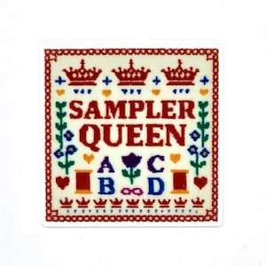 May include: A square sticker with a cross-stitch design. The sticker features the words "SAMPLER QUEEN" in red letters, surrounded by crowns, flowers, and spools of thread. The border is a red and cream color.
