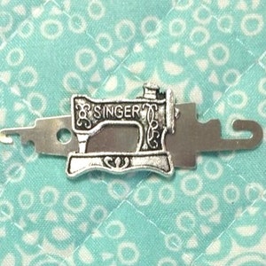 May include: A silver sewing machine-shaped threader tool with a hook on one end and a loop on the other. The tool is designed to help thread needles easily.