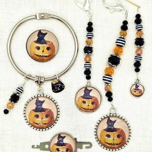 May include: A collection of Halloween-themed accessories featuring black cats and jack-o'-lanterns. The set includes a bracelet, keychains, and earrings. The charms have a black cat sitting inside an orange pumpkin. The keychains and earrings have black, orange, and white beads.