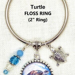 K&ouml;nnte beinhalten: Ein silberner Zahnseidenring mit einem Durchmesser von etwa 5 cm und einem Schildkr&ouml;ten-Design. Er enth&auml;lt einen Anh&auml;nger mit einem Schildkr&ouml;tenbild, einen kleinen Schildkr&ouml;ten-Charm und blaue Perlenakzente. Der Text "Turtle FLOSS RING (2" Ring)" ist abgebildet.