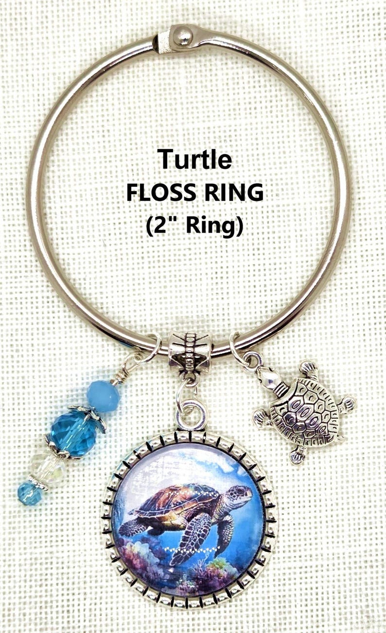 K&ouml;nnte beinhalten: Ein silberner Zahnseidenring mit einem Durchmesser von etwa 5 cm und einem Schildkr&ouml;ten-Design. Er enth&auml;lt einen Anh&auml;nger mit einem Schildkr&ouml;tenbild, einen kleinen Schildkr&ouml;ten-Charm und blaue Perlenakzente. Der Text "Turtle FLOSS RING (2" Ring)" ist abgebildet.