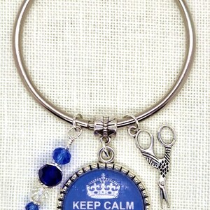 Puede incluir: Un llavero plateado con un dije azul y plateado. El dije tiene el texto "KEEP CALM AND Stitch On!" y una corona. Un dije de tijeras y cuentas azules completan el dise&ntilde;o. El llavero es probablemente para un artesano.