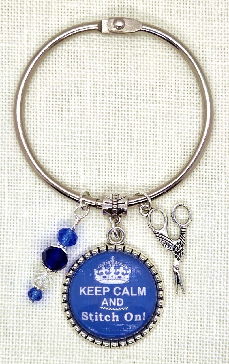 Puede incluir: Un llavero plateado con un dije azul y plateado. El dije tiene el texto "KEEP CALM AND Stitch On!" y una corona. Un dije de tijeras y cuentas azules completan el dise&ntilde;o. El llavero es probablemente para un artesano.
