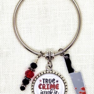 May include: A silver keychain with a round pendant that says "TRUE CRIME junkie" and a small knife charm with red blood splatter. The keychain also has red and black beads.